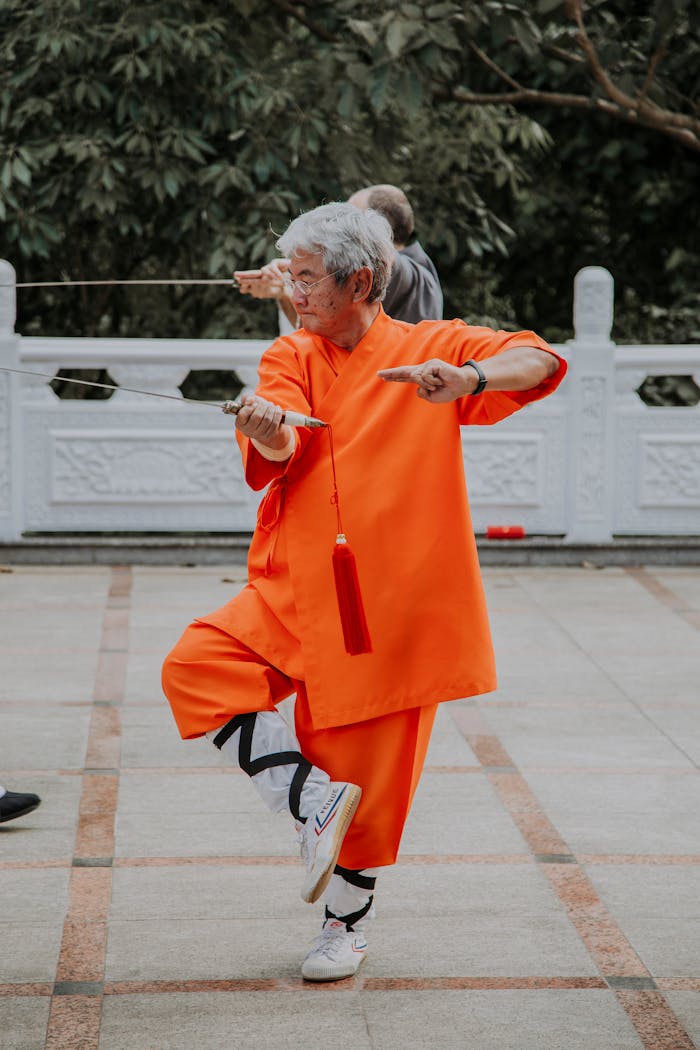 Senior ethnic male in orange apparel standing on one leg with tai chi sword on pavement while looking away
