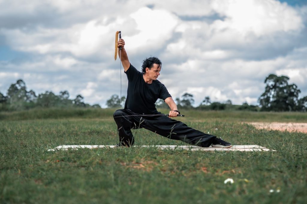 A martial artist practicing sword techniques in a scenic grassy field under a cloudy sky.